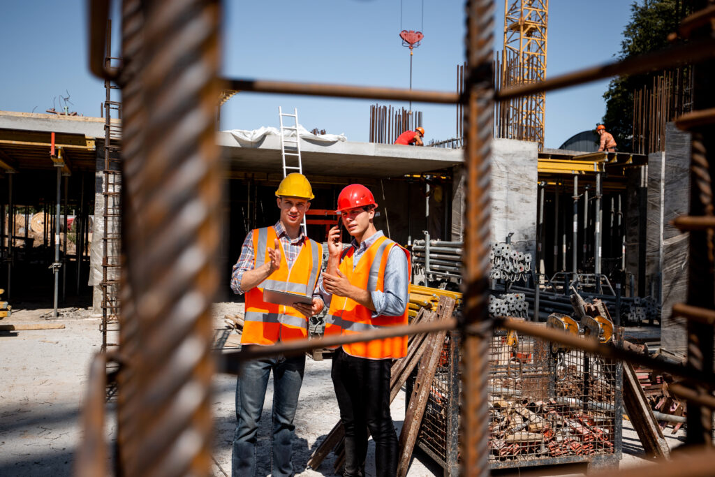 Architect and structural engineer in orange work vests and helmets discuss a building project on the open air building site with a lot of steel frames . .